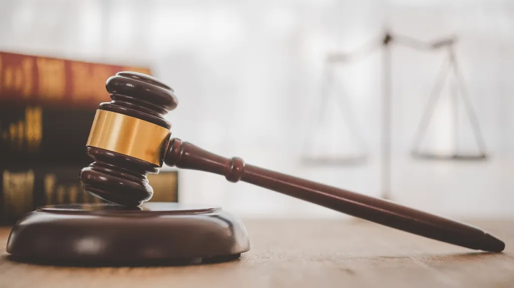 Wooden judge's gavel with brass band resting on sound block, law books and scales in background.