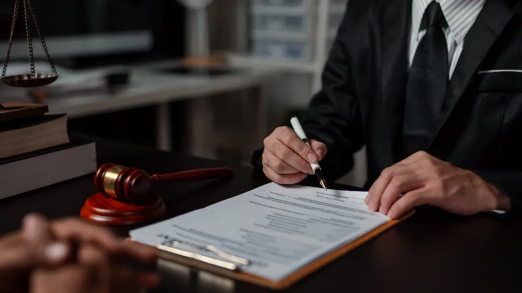 Person in formal suit reviewing and signing legal documents at desk with gavel and scales.
