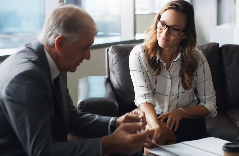 Two professionals, a man in a suit and a woman in glasses, discuss documents on a couch.