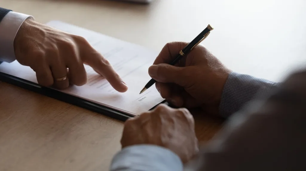 Two individuals reviewing and signing a document on a wooden table, one pointing at the signature line.