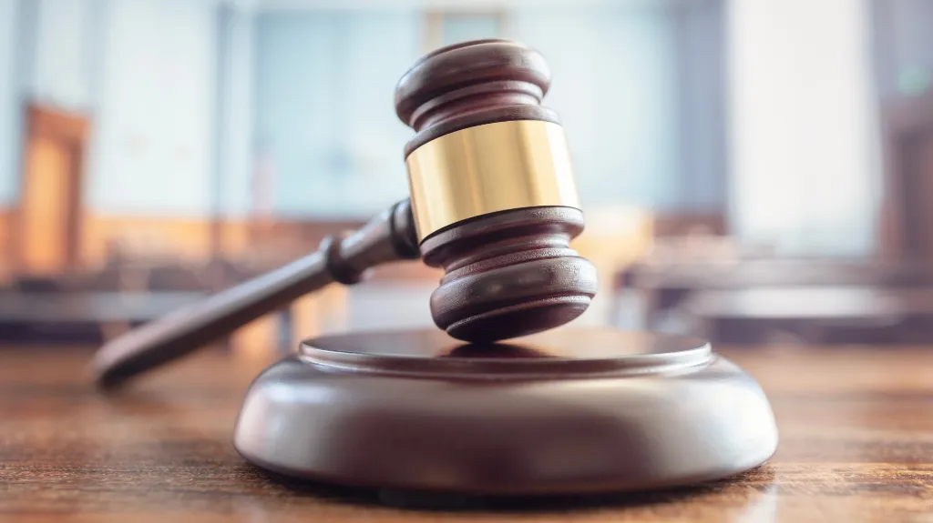 Wooden judge's gavel with gold band resting on sounding block in courtroom setting.