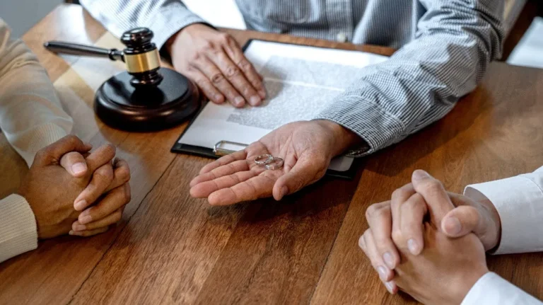 Two individuals with clasped hands across a wooden table from a person holding wedding rings and a gavel.
