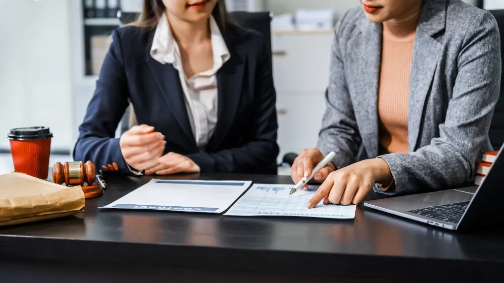 Two professional women reviewing documents at a desk with a laptop and gavel nearby.