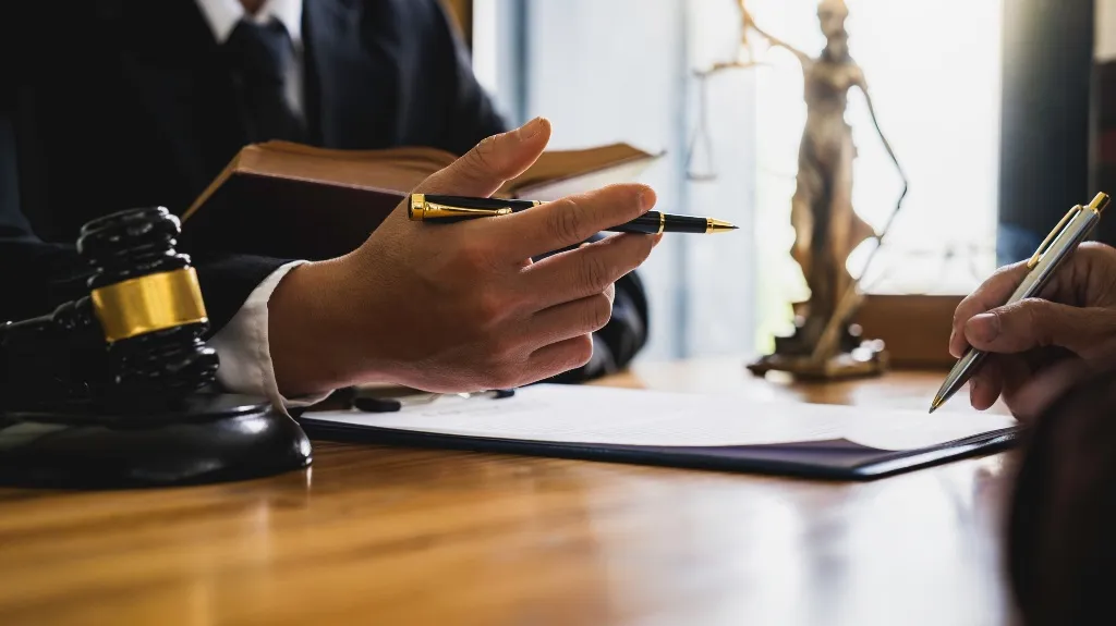 Two individuals in formal attire discussing documents at a wooden desk with a gavel and Lady Justice statue.