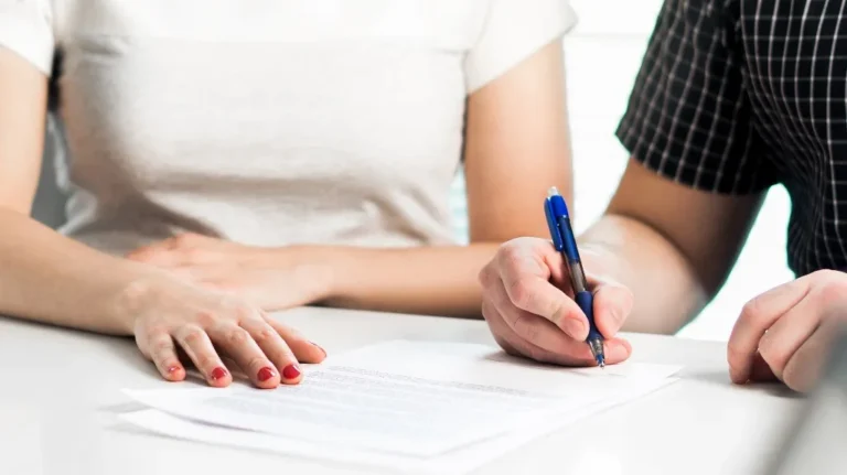 Two individuals at a table, one signing a document with a blue pen, the other resting hands nearby.