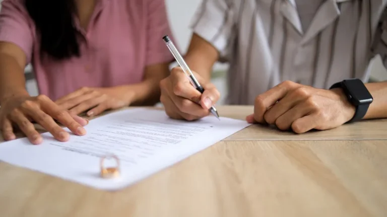 Two people signing a document on a wooden table, with a ring placed nearby.
