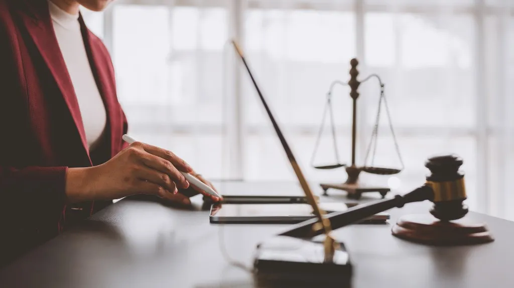 Person in a burgundy blazer using a stylus on a tablet, with scales of justice and gavel on desk.