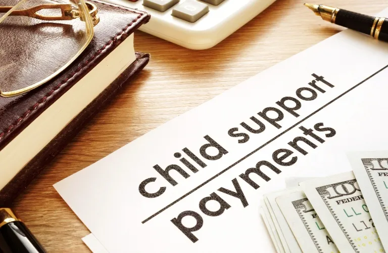 Document titled "child support payments" on a wooden desk with currency, eyeglasses, notebook, pen, and calculator.