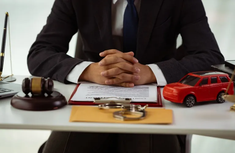 Person in dark suit sitting at desk with gavel, handcuffs, toy car, and legal documents.