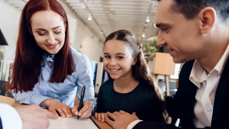 Woman signing a document at a table with a smiling young girl and a man in a business setting.
