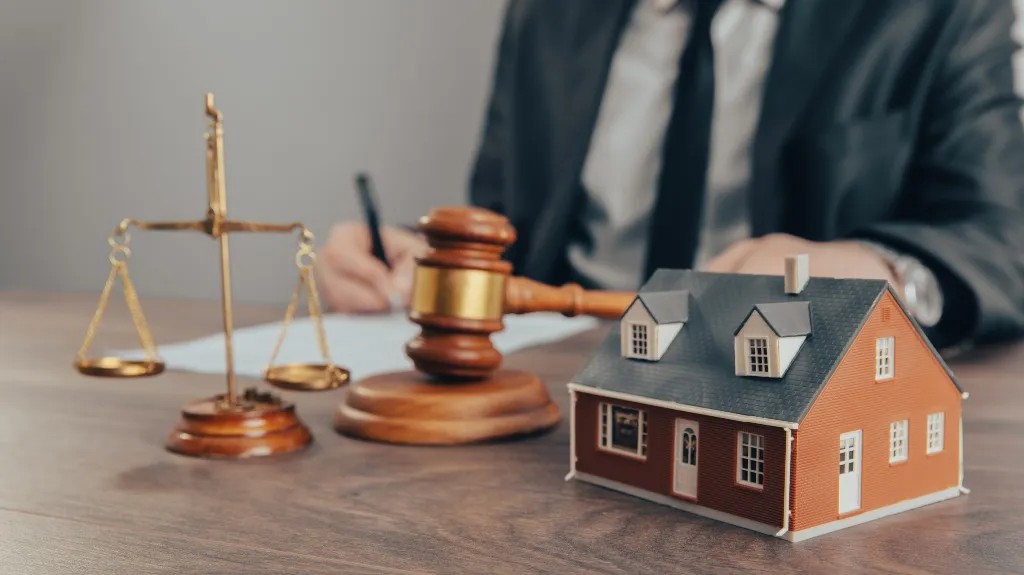 Scale model of a house, wooden gavel, and brass scales of justice on desk.