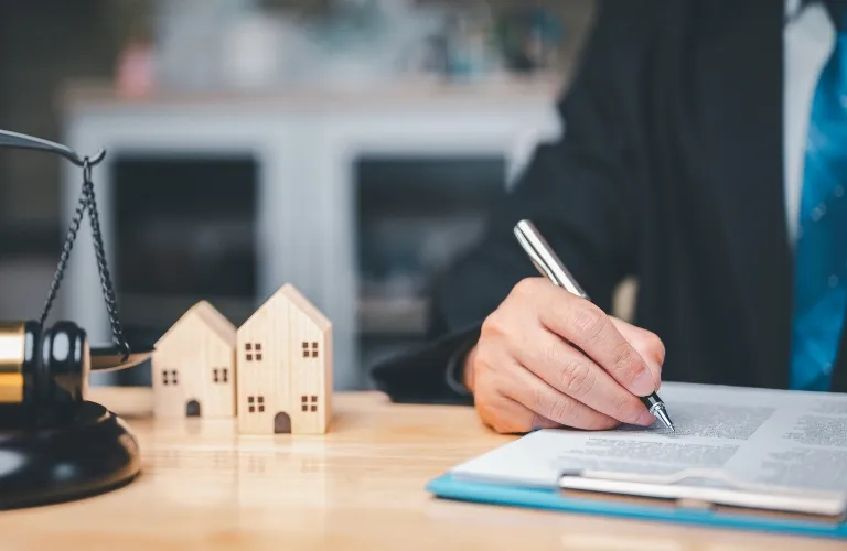 Person in business attire signing legal documents on a desk with wooden house models and scales of justice.