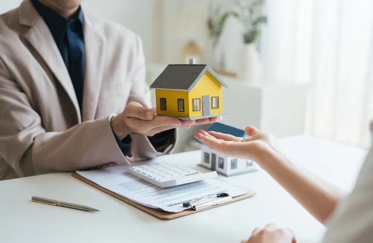 Person in beige suit holding yellow house model towards another person's outstretched hand over desk with documents and calculator.