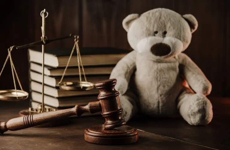 Wooden judge's gavel, brass balance scales, legal books, and a beige teddy bear on a wooden surface.