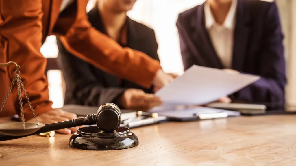 Wooden gavel and scales of justice on table with two business professionals reviewing documents.