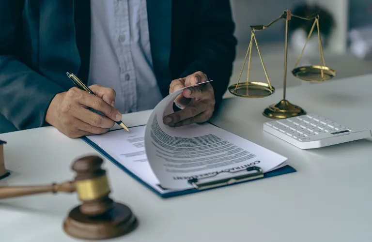 Person in formal attire reviewing and signing legal documents with gavel and scales of justice on desk.