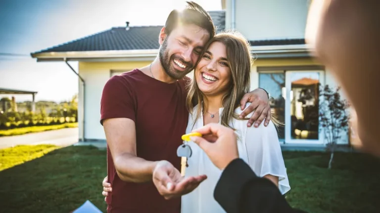 Smiling couple receiving house keys outdoors in front of modern home during daytime.
