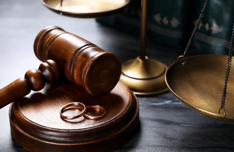 Wooden judge's gavel, two gold wedding rings, and brass scales of justice on dark surface.