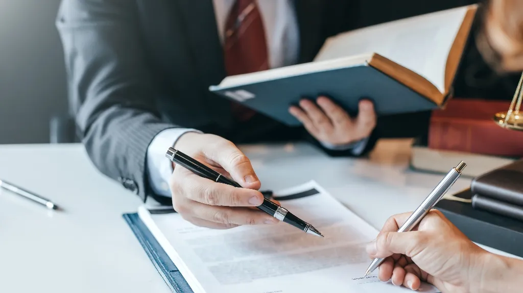 Two individuals in formal attire reviewing and signing a document at a desk.