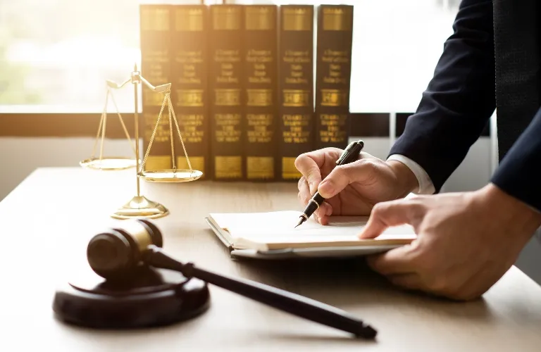 Person in dark suit writing on document at desk with legal books, scales of justice, and gavel.