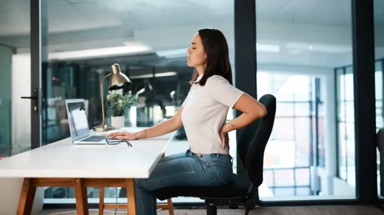 Young woman in white shirt and jeans sitting at desk, holding lower back in discomfort.