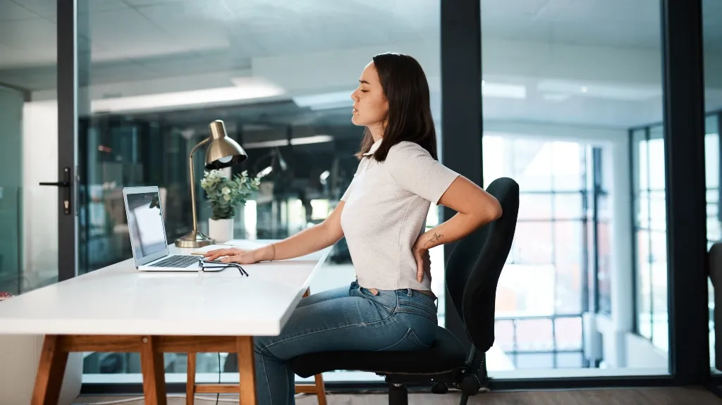 Young woman in white shirt and jeans sitting at desk, holding lower back in discomfort.