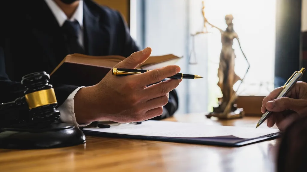 Two professionals in formal attire discussing documents, with gavel and Lady Justice statue on desk.